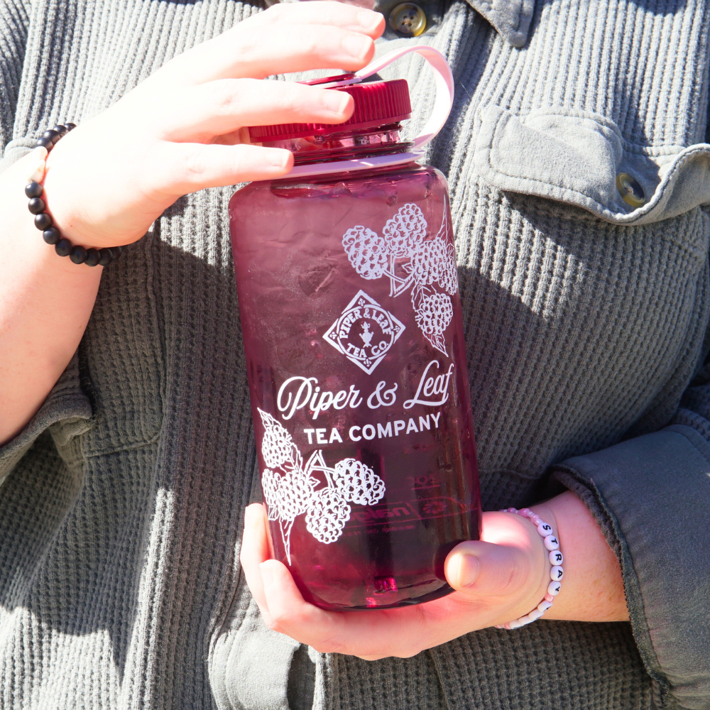 A close up of a woman holding a large translucent 32oz Wide Mouth Piper & Leaf Nalgene Water (Tea) Bottle - Into The Briar Patch, filled with a dark beverage, labeled "Piper & Leaf Tea Co." in front of her.