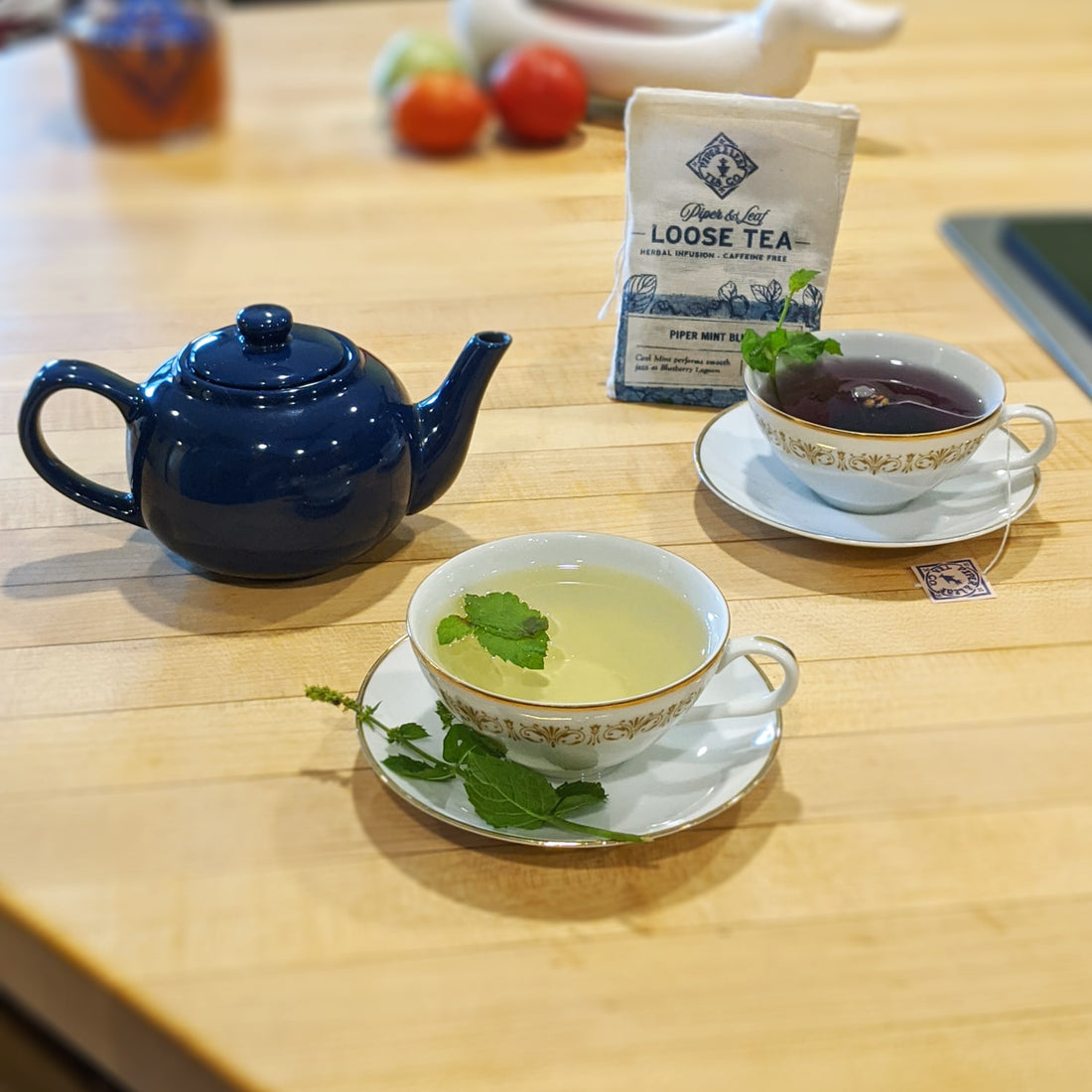 Two teacups: one of fresh mint tea and the other full of a blue herbal tea. A bag of Piper Mint Blues sachets sits in the background