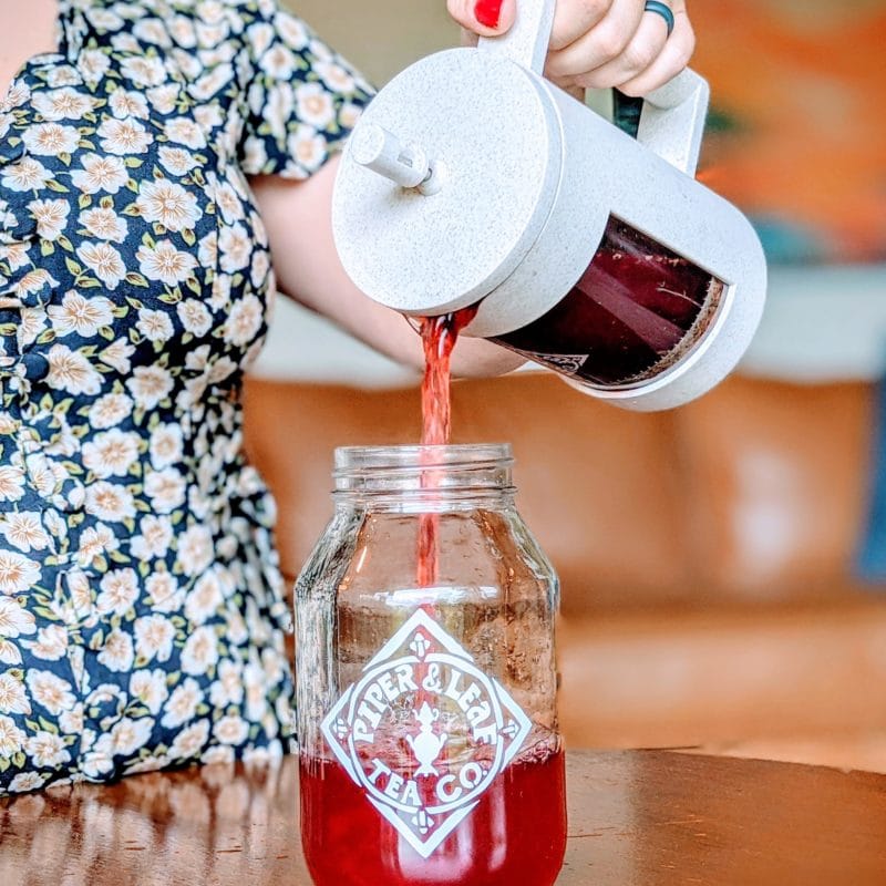 A person pours red tea from a French press into a glass jar labeled "Piper & Leaf Tea Co." on a wooden surface.