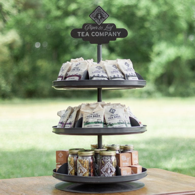 Three-tiered display stand holding Piper & Leaf Tea Company tea bags on the top two tiers and jars of loose tea on the bottom tier, set outdoors on a wooden table.