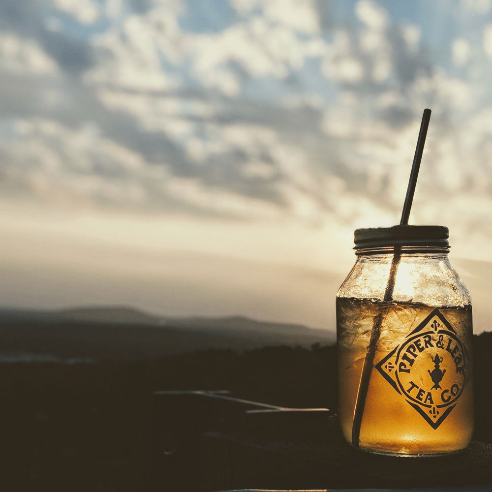 A jar of iced tea with a straw sits on a surface outdoors, with a mountain landscape and cloudy sky in the background at sunset.
