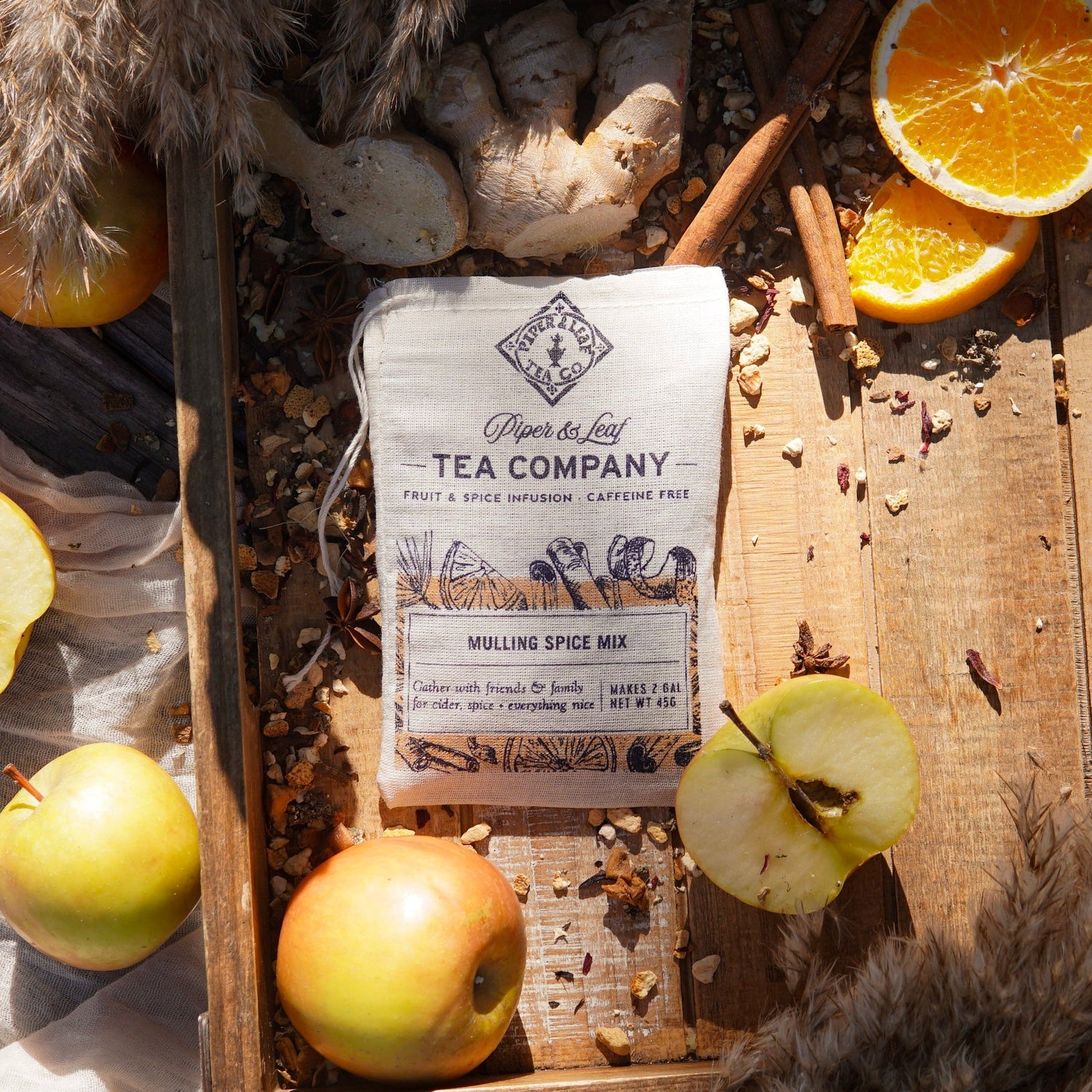 A cloth bag labeled "Piper & Leaf Tea Company Mulling Spice Mix" sits between two red apples on a brown surface.