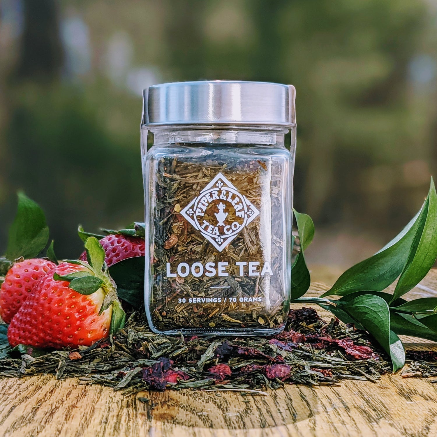A jar of River & Leaf Tea Co. loose tea surrounded by loose leaves, strawberries, and green foliage on a wooden surface.