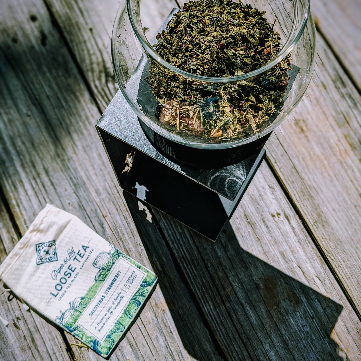 A glass bowl of loose tea leaves sits on a square black scale next to a white loose tea pouch, all placed on a weathered wooden surface.