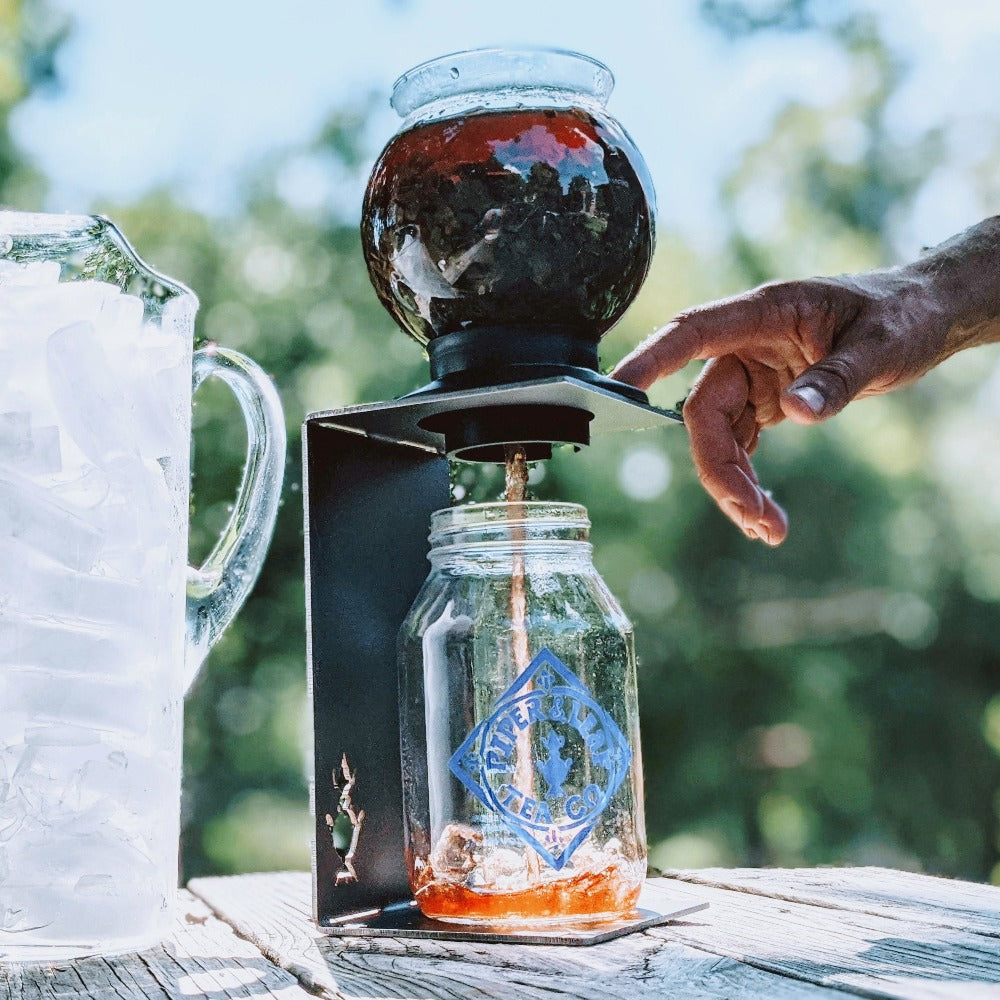 A person operates a cold brew coffee maker, with coffee dripping into a glass jar beside a pitcher filled with ice on a wooden table outdoors.