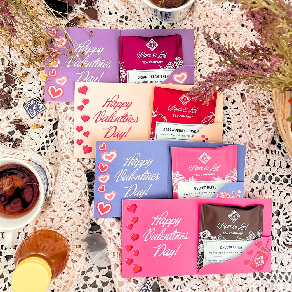 Valentine’s Day cards and assorted Piper & Leaf tea packets are arranged on a lace tablecloth with cups of tea and decorative flowers nearby.