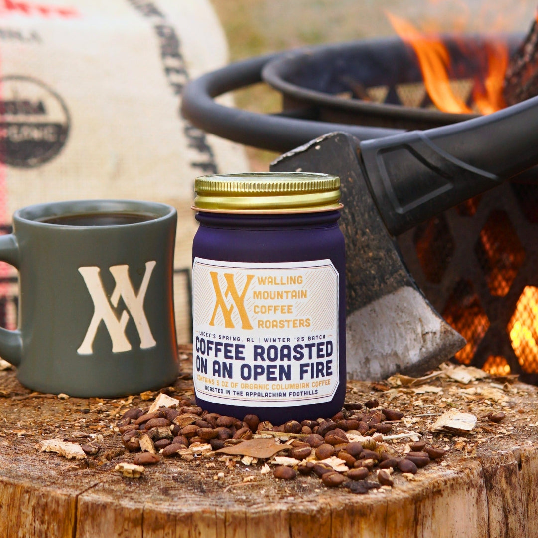 Coffee bags and a mug with a logo sit on a wooden surface surrounded by evergreen branches, with blurred trees and string lights in the background.