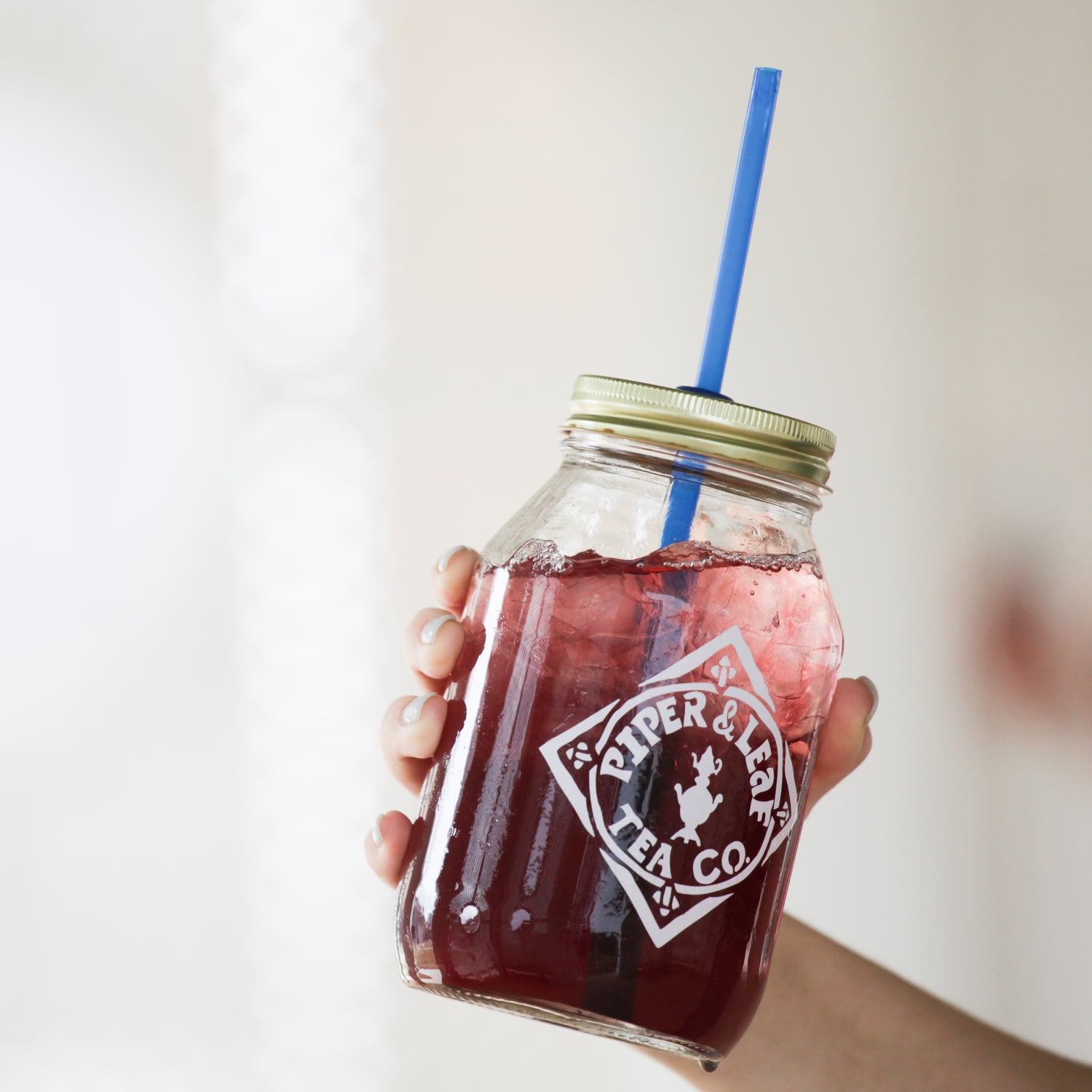 A hand holding a glass jar with a metal lid filled with iced red tea and a blue straw. The jar is labeled "Piper & Leaf Tea Co.
