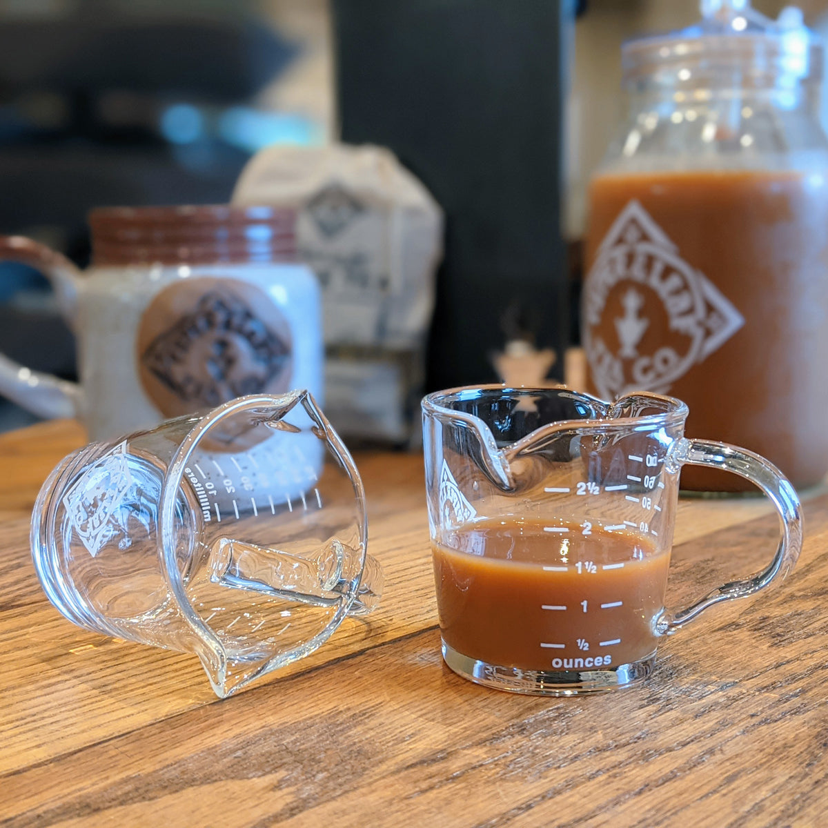 Two glass measuring cups are on a wooden table, one tipped over and empty, the other upright and filled with coffee. Bags and jars are in the background.