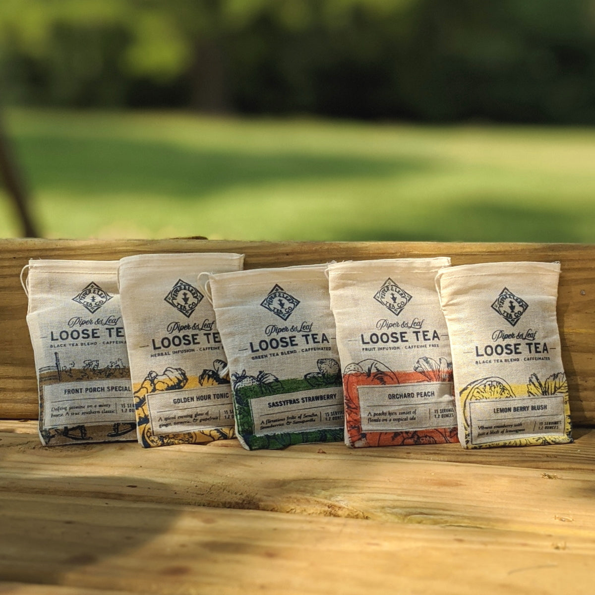 Five bags of loose leaf tea labeled with different flavors are displayed on a wooden surface outdoors with greenery in the background.