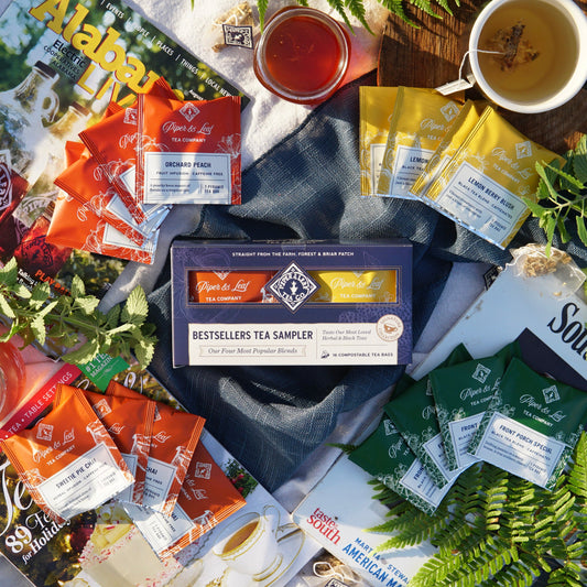 Piper & Leaf Tea Co.'s Bestsellers Tea Sampler, a box of 16 tea bags with unique herbal blends, is displayed on magazines and ferns next to a steaming cup of tea.
