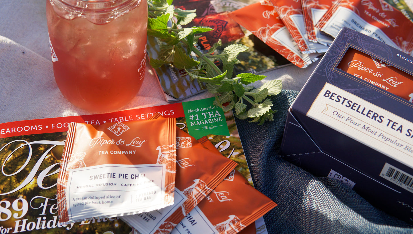 Piper & Leaf Tea Co.’s Bestsellers Tea Sampler (16 tea bags), assorted tea packets, a glass of iced tea, fresh mint, and a tea-themed magazine are displayed on the table.