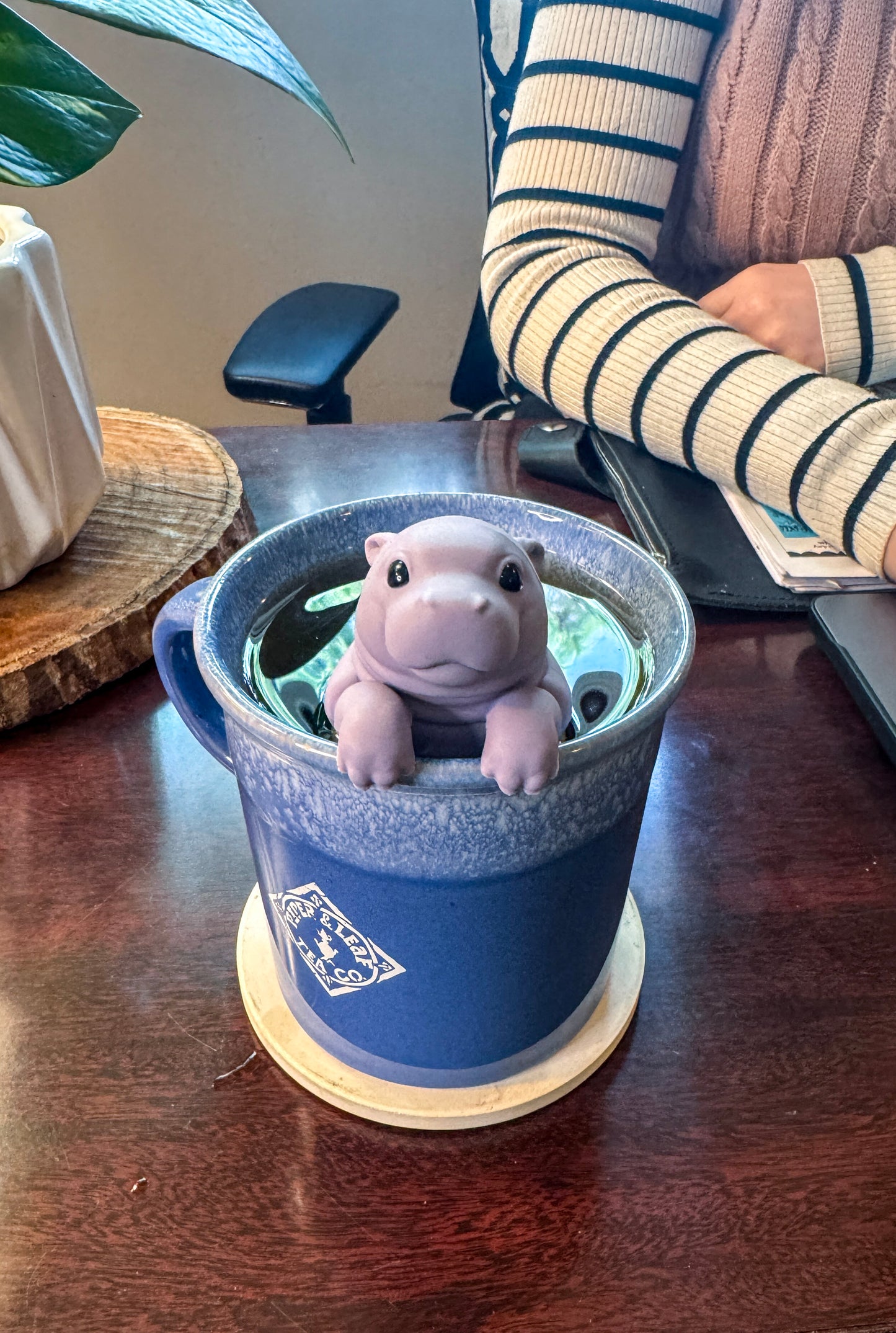 The Piper & Leaf Tea Co. Sip'Opotamus Infuser, a small BPA-free hippopotamus-shaped tea infuser, sits in a blue mug on a wooden desk near a person in a striped sweater.
