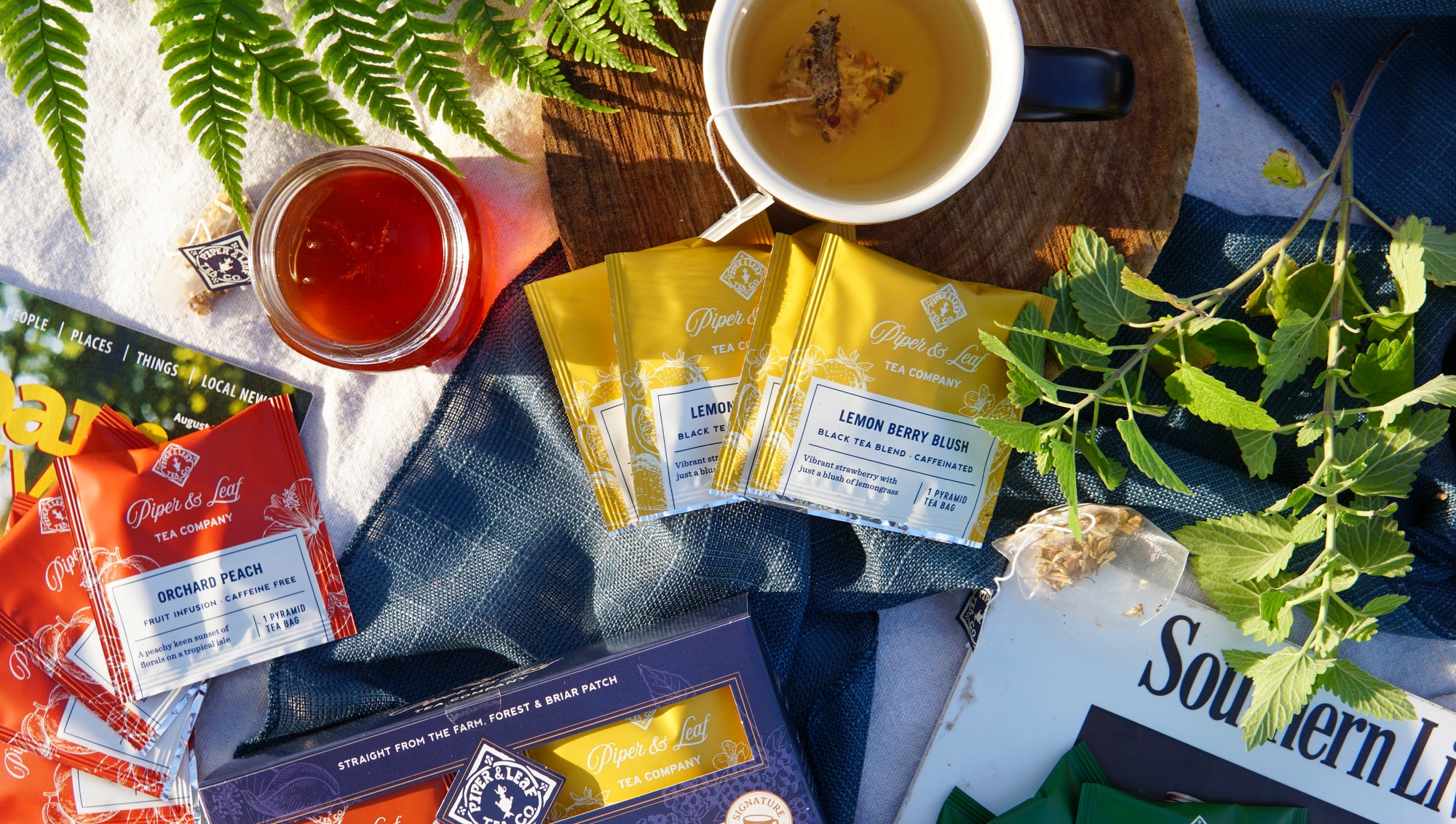 A box of 16 Piper & Leaf Tea Co. Bestselling Tea Sampler bags, a cup of black tea blend, an open honey jar, a plant, and the book "Southern" are arranged on a blue and brown surface.