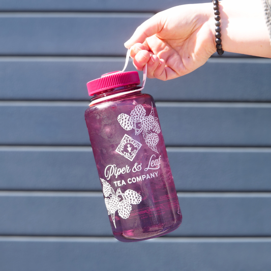A hand holding a large translucent 32oz Wide Mouth Piper & Leaf Nalgene Water (Tea) Bottle - Into The Briar Patch, filled with a dark beverage, labeled "Piper & Leaf Tea Co." The background is a dark blue garage door 