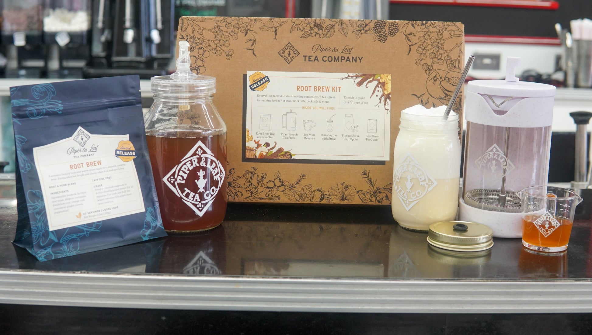 A Piper and Leaf Tea Co. Root Beer Tea Brew Kit sits on a counter, featuring a tea package, tea press, concentrated tea jar, mason jar with powder, pitcher, small glass of liquid, and a branded box.