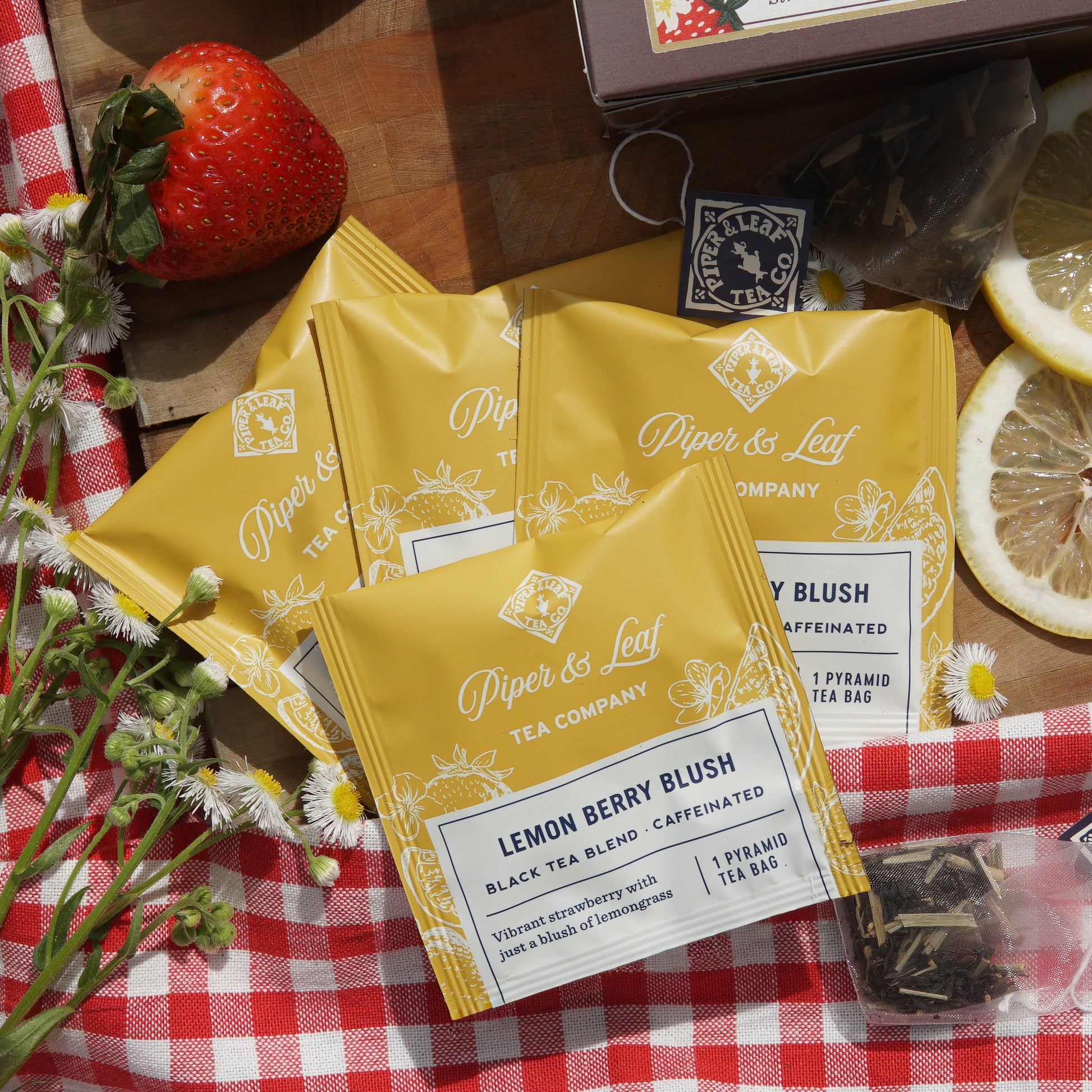 Lemon Berry Blush indiviually wrapped tea bags on top of a picnic blanket and wooden cutting board, surrounded by assorted lemons, strawberries, and wild flowers