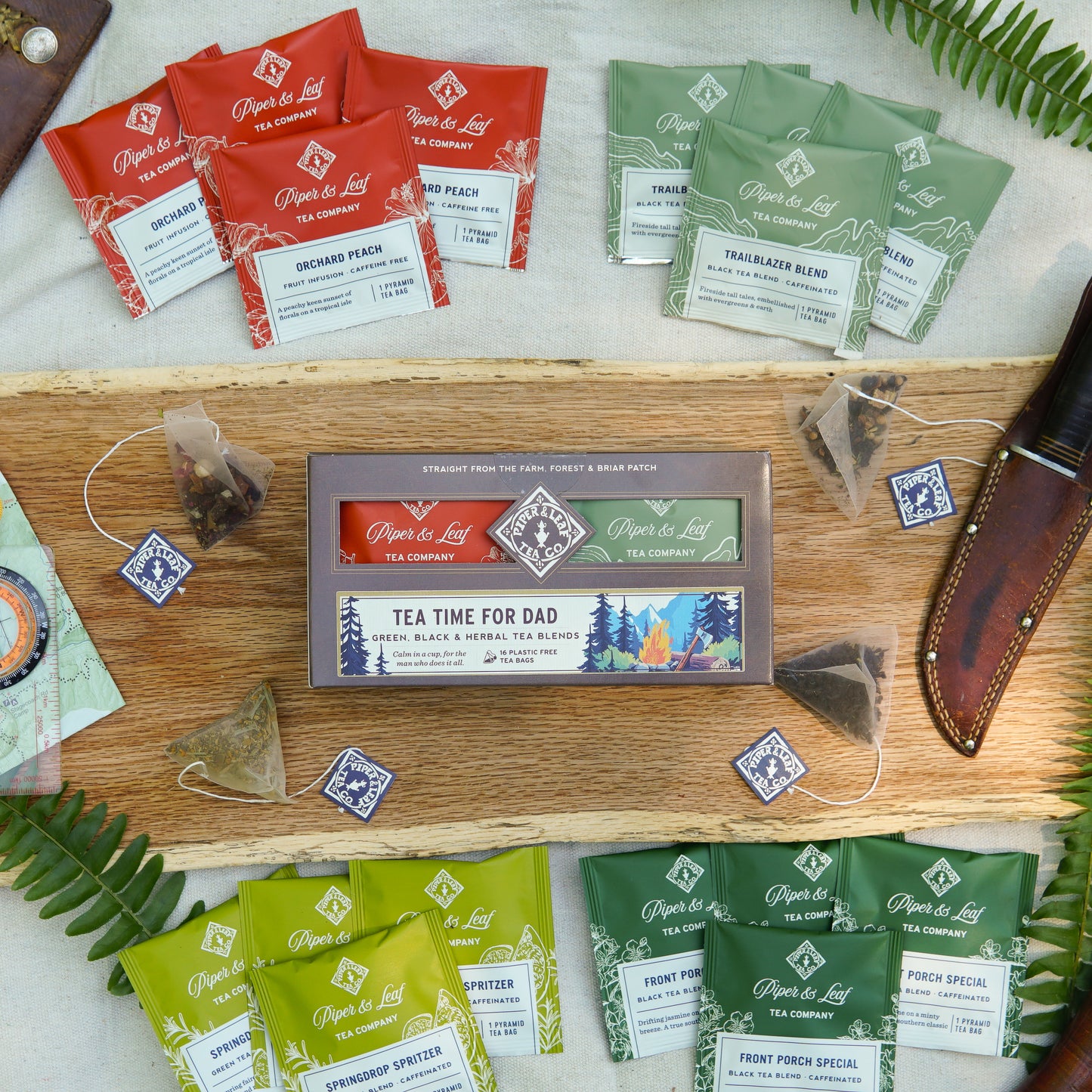 A Piper & Leaf Tea Co. “Tea Time For Dad” box with 16 individually wrapped tea bags is displayed on a table alongside other Piper & Leaf tea packets, tea bags, a knife, and fern leaves.