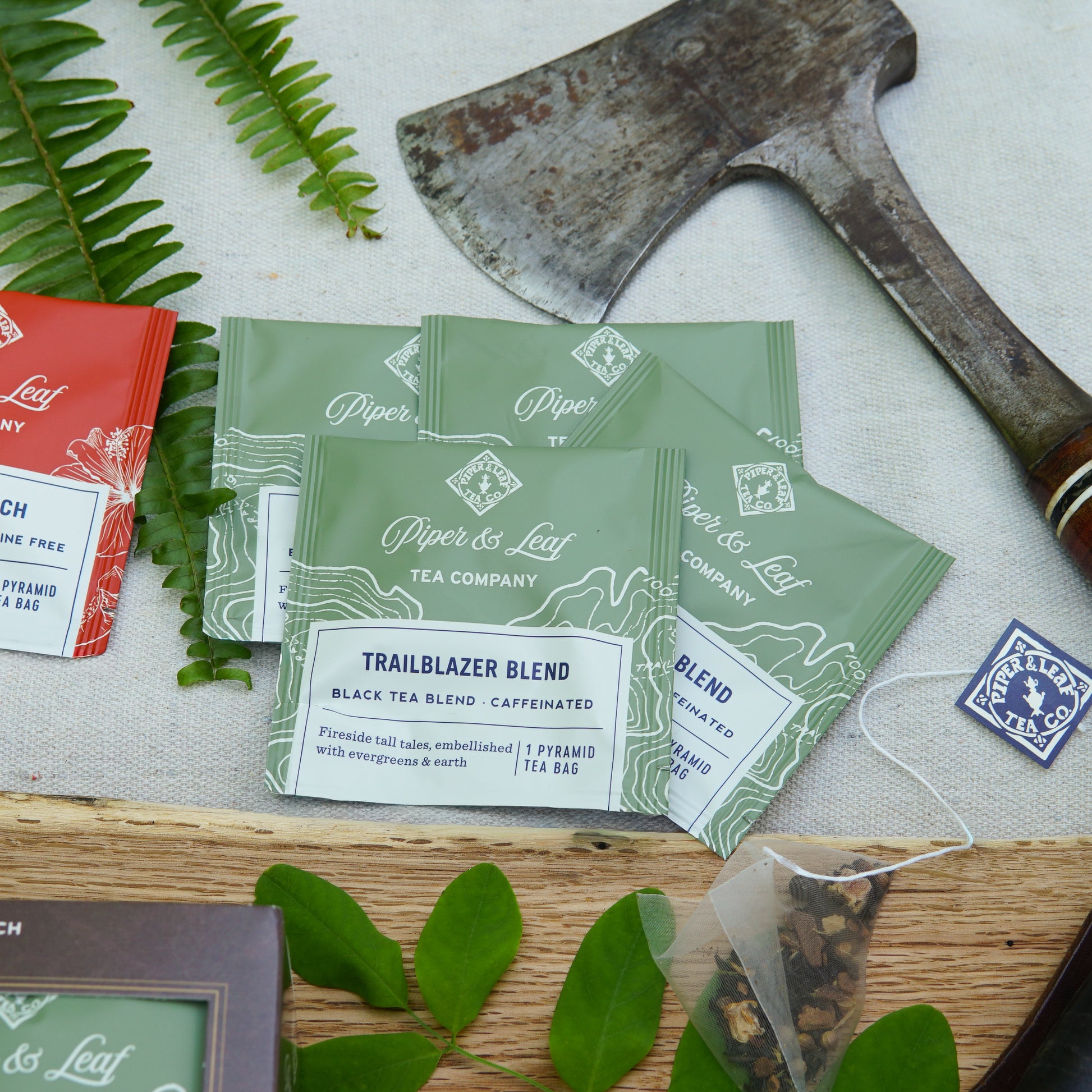 Several green Piper & Leaf Tea Co. "Tea Time For Dad" boxes, each with 16 individually wrapped tea bags, are arranged on a white cloth next to an axe and green leaves—making a unique curated Father’s Day tea gift. Other teas are partially shown.