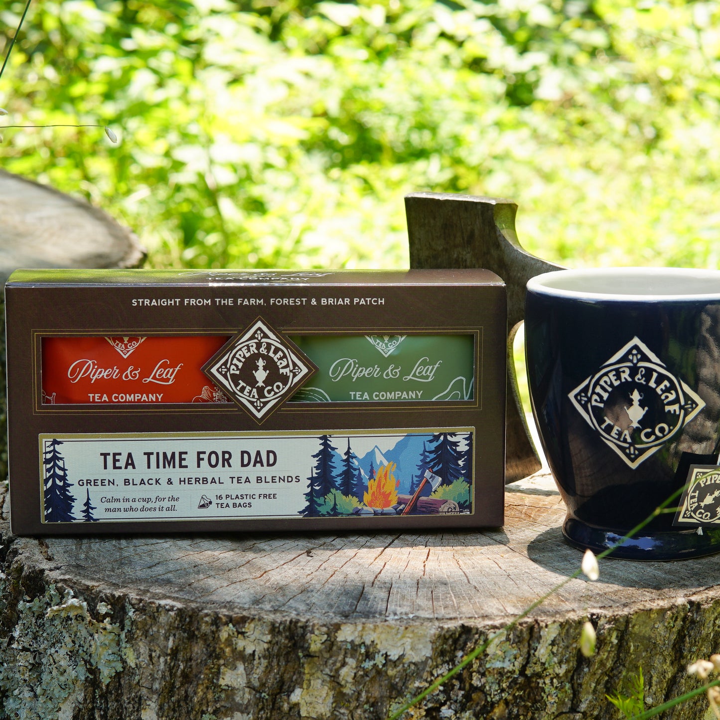 The "Tea Time For Dad- Box of 16 Individually Wrapped Tea Bags" from Piper & Leaf Tea Co. is pictured on a tree stump beside a navy blue mug, with an axe nearby—an ideal Father’s Day tea gift with rustic charm.