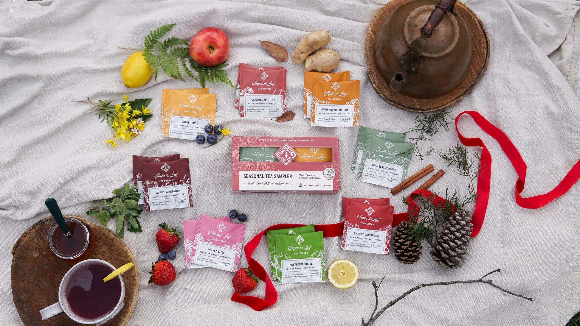 The Piper & Leaf Tea Co. Seasonal Tea Sampler (Box of 16 Tea Bags) is beautifully arranged with fruit, flowers, pinecones, a red ribbon, ceramic teapot, and cup—an ideal seasonal tea box display.
