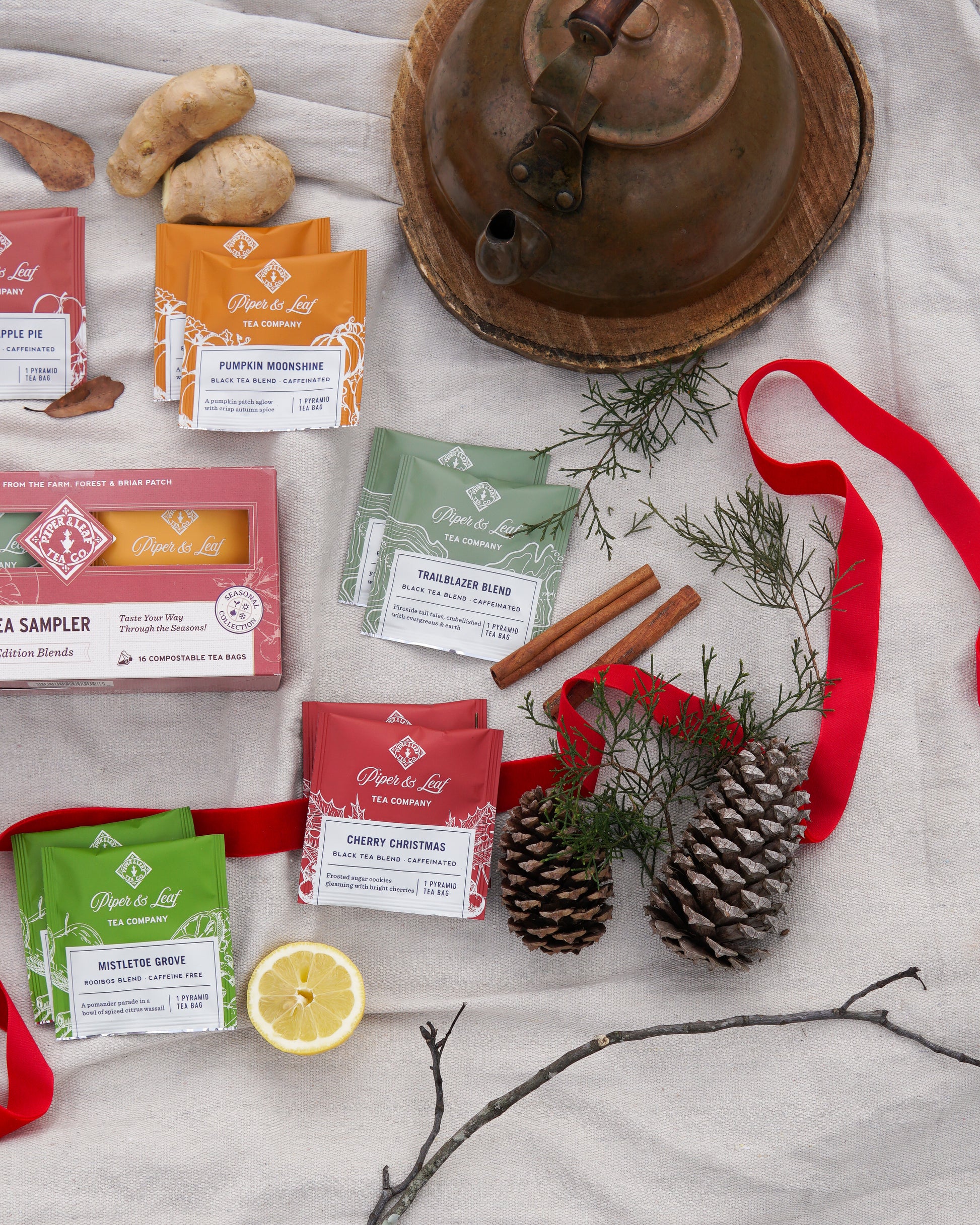 The Piper & Leaf Tea Co. Seasonal Tea Sampler (Box of 16 Tea Bags) is displayed with pinecones, cinnamon sticks, lemon slices, a teapot on wood, red ribbon, greenery, and ginger pieces on light fabric.