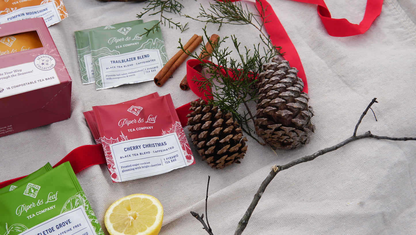 The Piper & Leaf Tea Co. Seasonal Tea Sampler – Box of 16 Tea Bags is artfully arranged with pinecones, cinnamon, lemon, ribbon, and foliage on beige cloth—perfect for a cozy seasonal tea box display.