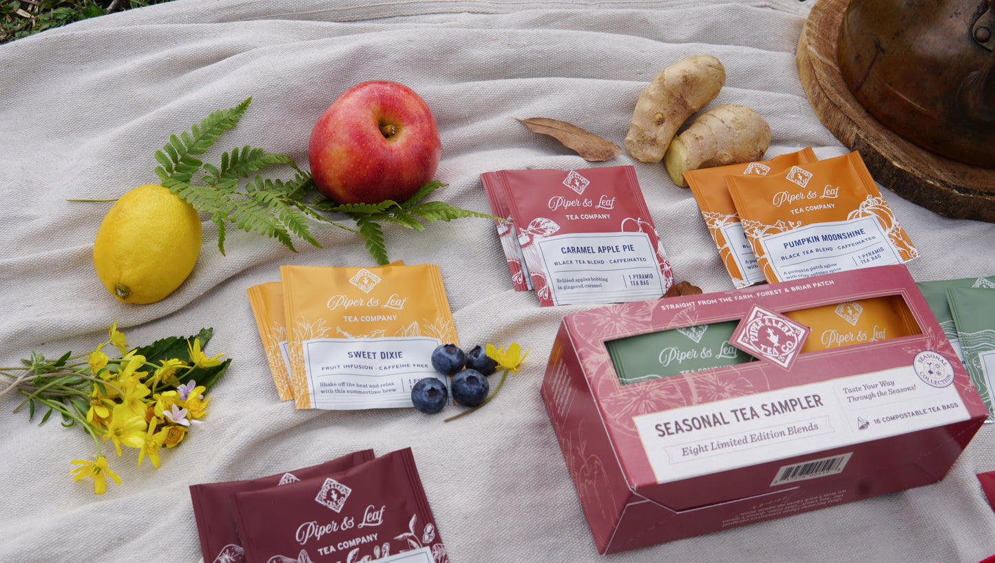 Piper & Leaf Tea Co.’s Seasonal Tea Sampler—Box of 16 Tea Bags—is artfully arranged with fruit, ginger, leaves, and flowers on a cloth outdoors.