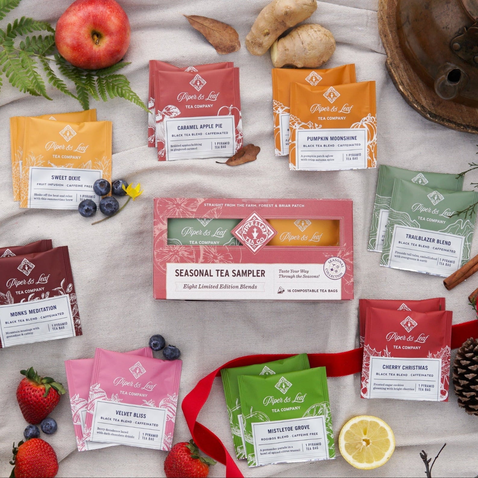 The Piper & Leaf Tea Co. Seasonal Tea Sampler—Box of 16 Tea Bags—is surrounded by eight assorted tea packets, fruit, cinnamon sticks, and a red ribbon, all artfully arranged on a white surface.
