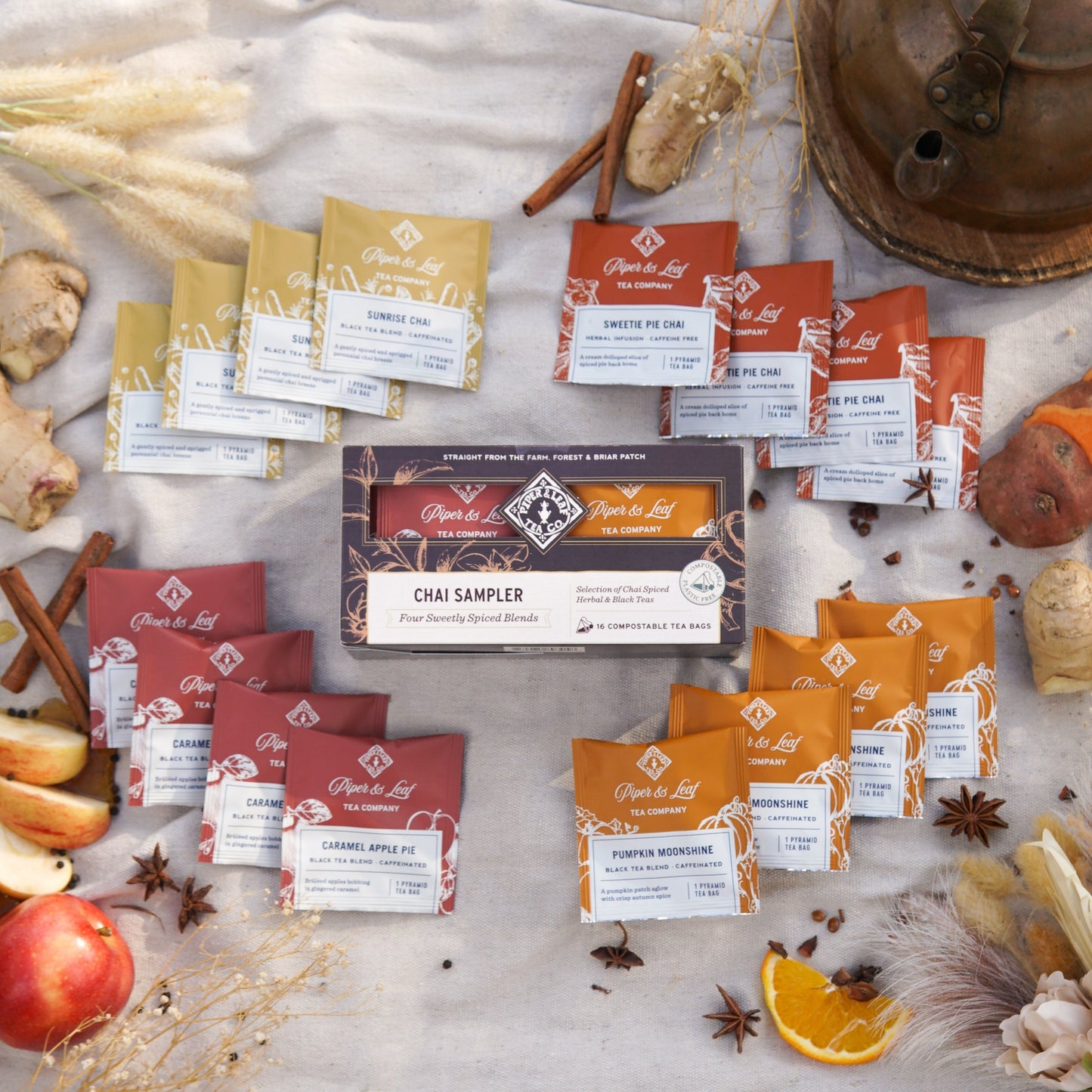 Sixteen Piper & Leaf Tea Co. chai sampler bags are artfully arranged around a fall-themed box with ginger, apple and orange slices, plus spices on a beige background—an inviting way to sample cozy autumn blends.