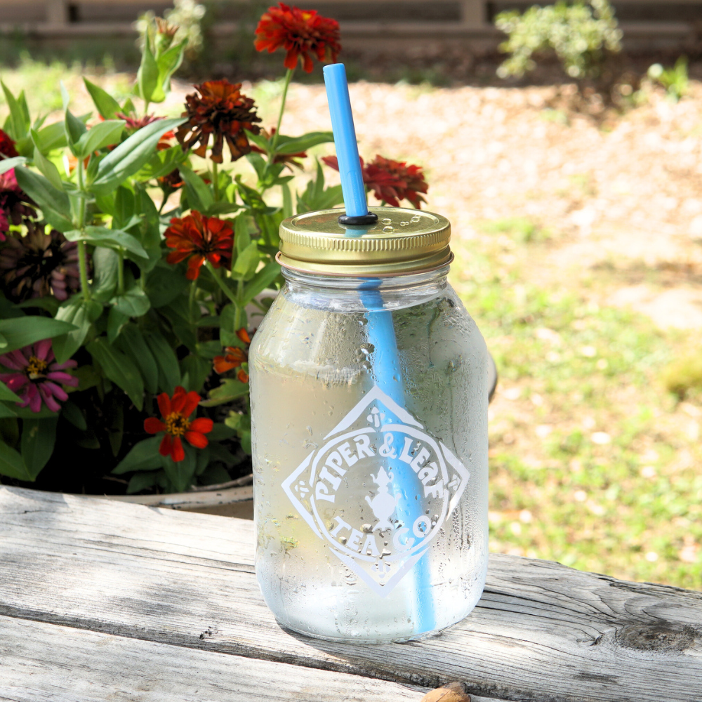 An UnStraw mason jar from Piper & Leaf Tea Co., featuring a gold lid and a blue straw, is positioned on a wooden table filled with iced water, next to vibrant potted red and orange flowers.