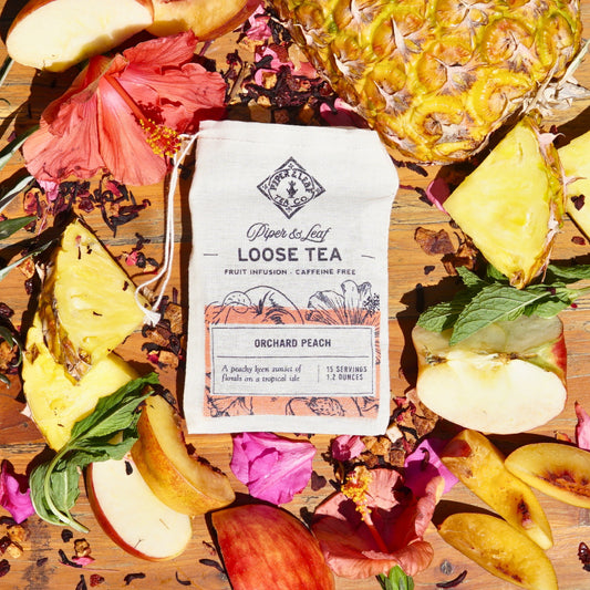 An Orchard Peach muslin bag from Piper & Leaf Tea Co., containing 15 servings of loose leaf tea, rests among sliced apples, pineapple pieces, hibiscus flowers, and lush tea leaves on a wooden surface.