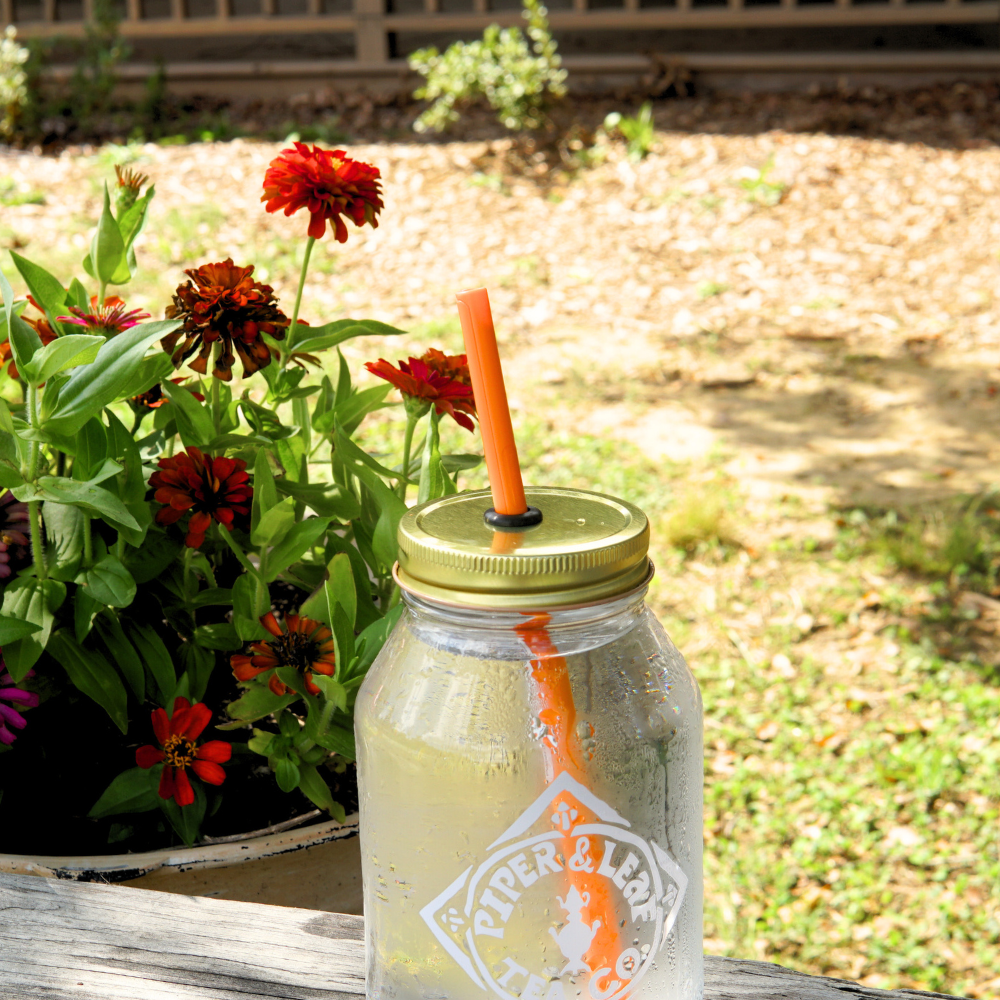 An UnStraw filled with a cold drink and an orange straw from Piper & Leaf Tea Co. sits on a wooden surface next to a pot of vibrant red flowers.