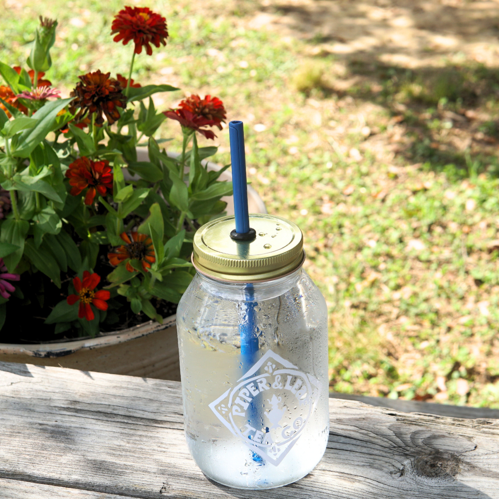 An UnStraw by Piper & Leaf Tea Co., featuring a glass jar filled with water, a golden lid, and a BPA-free blue straw, sits on a wooden bench with red and orange flowers in a pot blooming in the background.