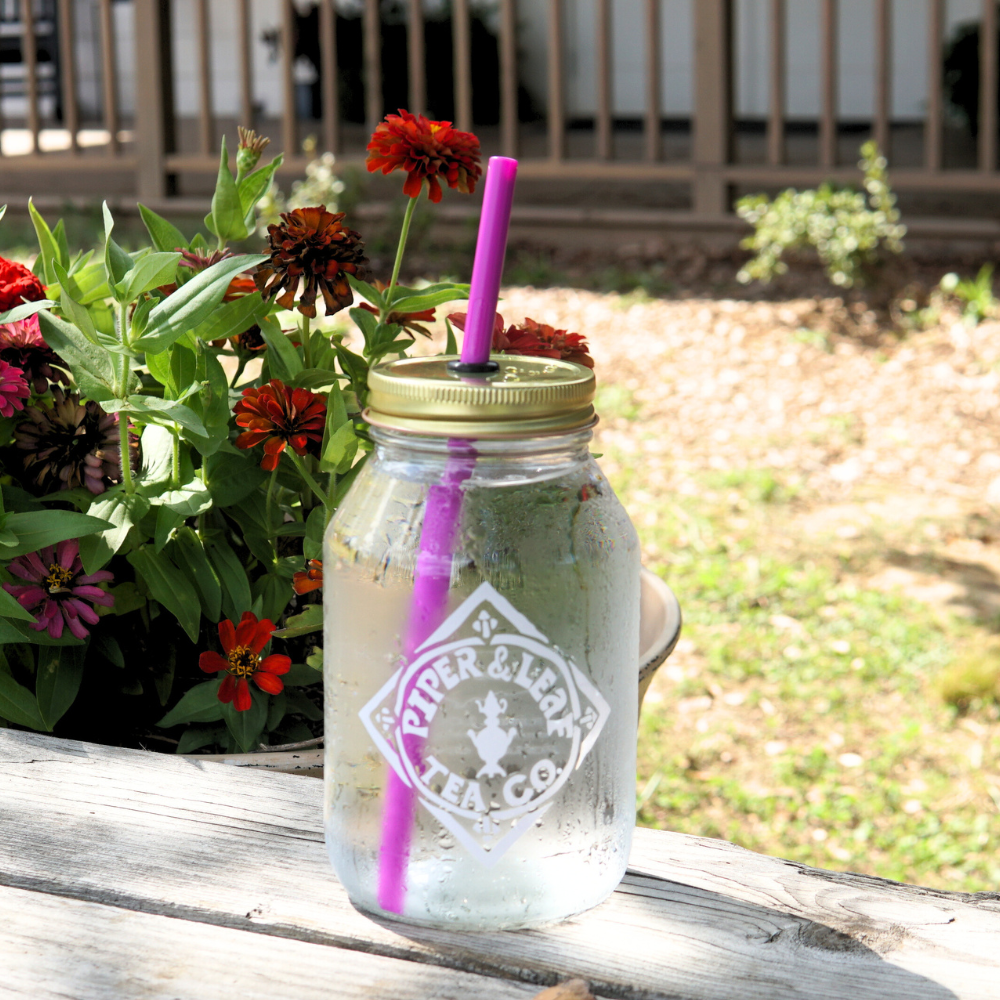 An UnStraw mason jar with a gold lid and purple straw, crafted with BPA-free materials, sits on a wooden surface near red flowers. The Piper & Leaf Tea Co. logo is prominently displayed on the jar, effortlessly blending style with safety.