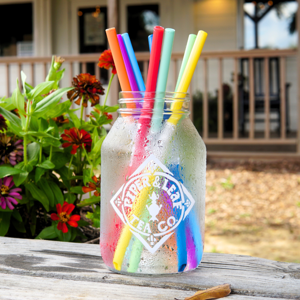 An UnStraw jar adorned with colorful, BPA-free straws from Piper & Leaf Tea Co. sits on a wooden table next to vibrant flowers.