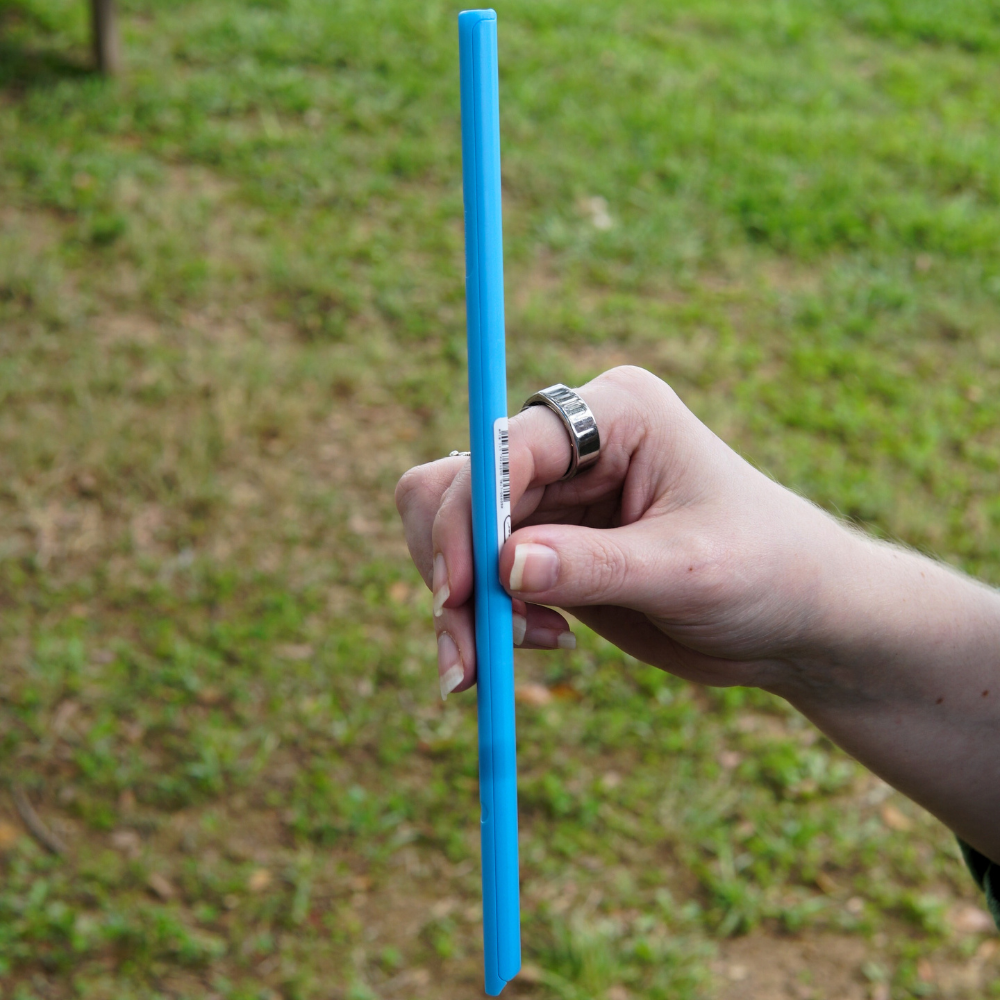A hand holds a large blue "UNSTRAW" from Piper & Leaf Tea Co. against a grassy background. This BPA-free straw boasts a split-apart design, making it both practical and vibrant.
