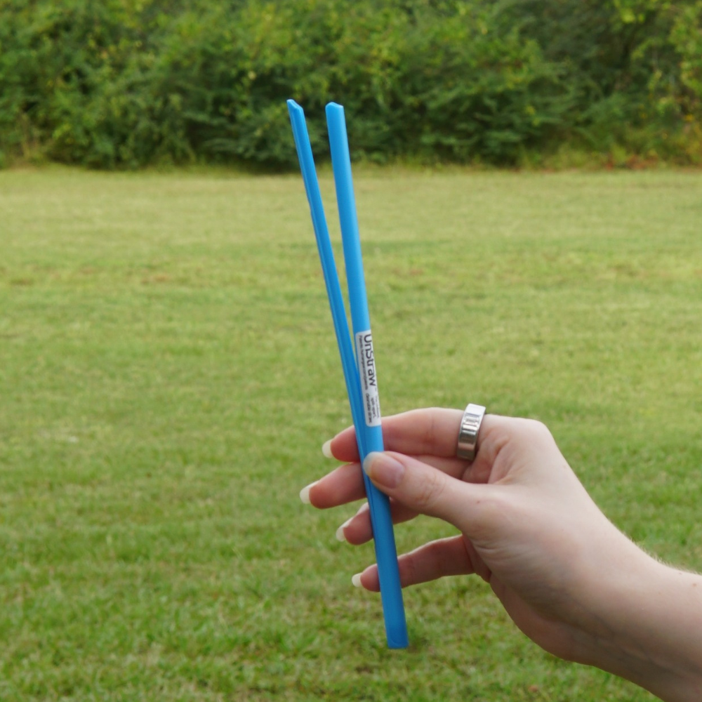 Someone holds a pair of Piper & Leaf Tea Co.'s UnStraw, two blue, BPA-free chopsticks, in front of a grassy field.