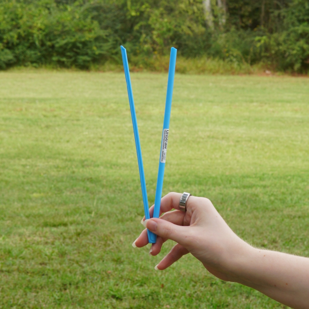 A hand holding two long, colorful UnStraws from Piper & Leaf Tea Co. in an outdoor grassy area, perfect for easy cleaning.