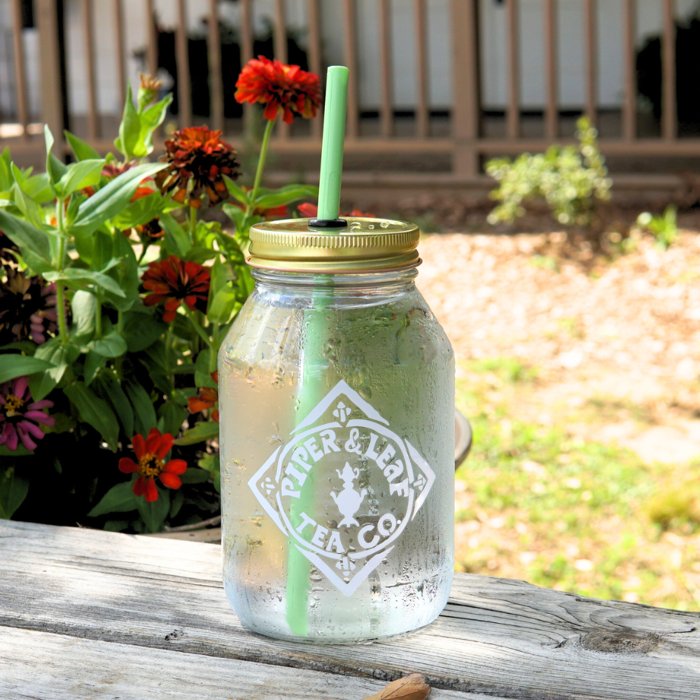 A Piper & Leaf Tea Co. glass jar filled with a chilled beverage and featuring an UnStraw, sits on a wooden surface next to vibrant red and orange flowers in a garden setting.