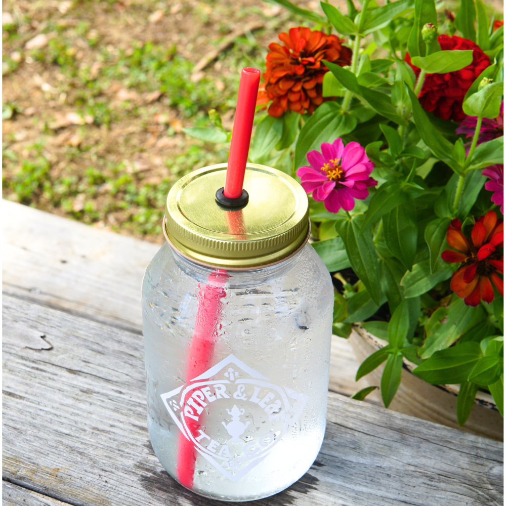 An UnStraw by Piper & Leaf Tea Co., featuring a golden lid and a BPA-free red straw, is filled with ice water and rests on a wooden bench beside vibrant flowers.