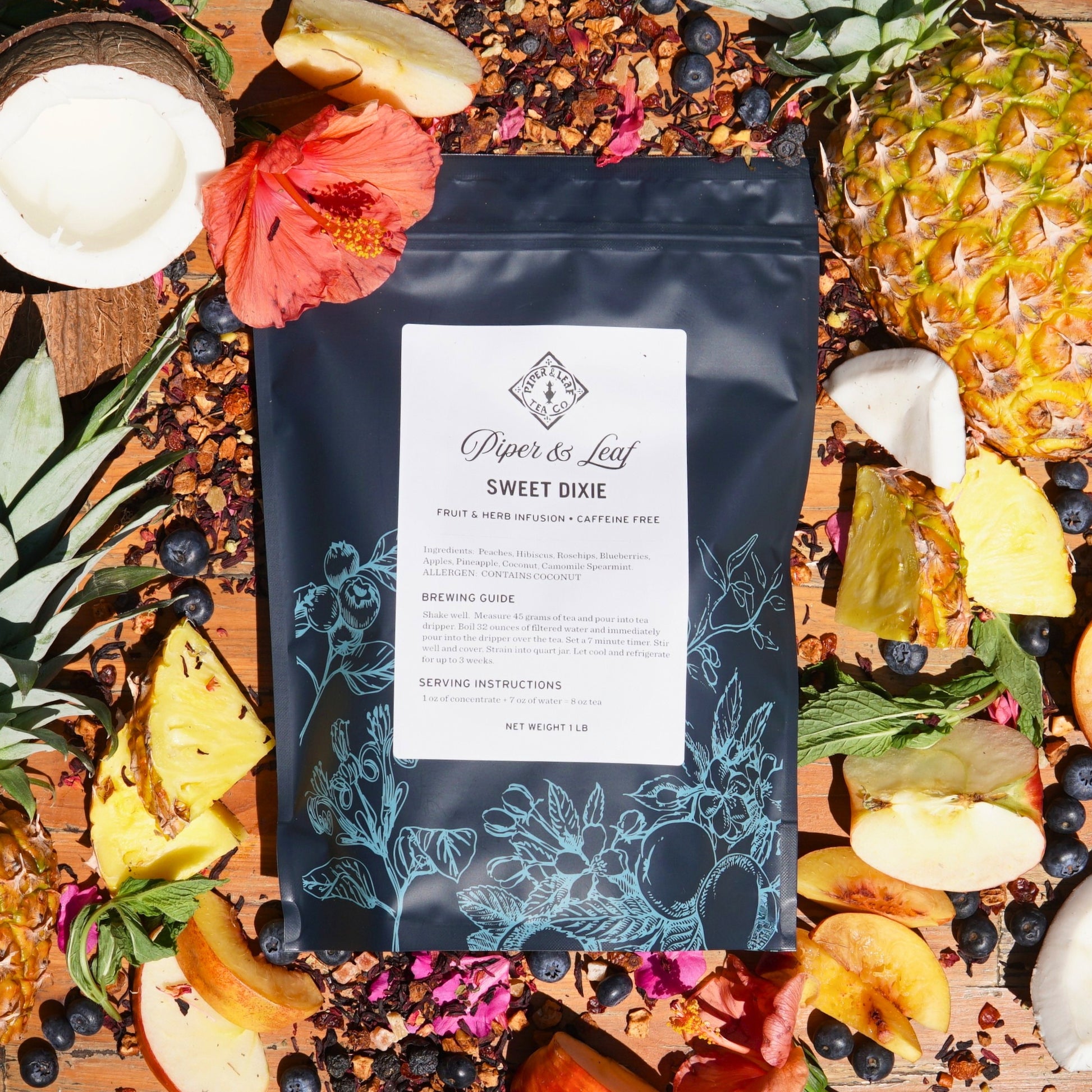 A bag of Piper & Leaf Tea Co.'s Sweet Dixie Pound Bag—190 servings—sits enveloped by a vibrant fruit medley with loose leaf tea, berries, pineapple, apples, and hibiscus on a wooden surface—a perfect summertime brew.