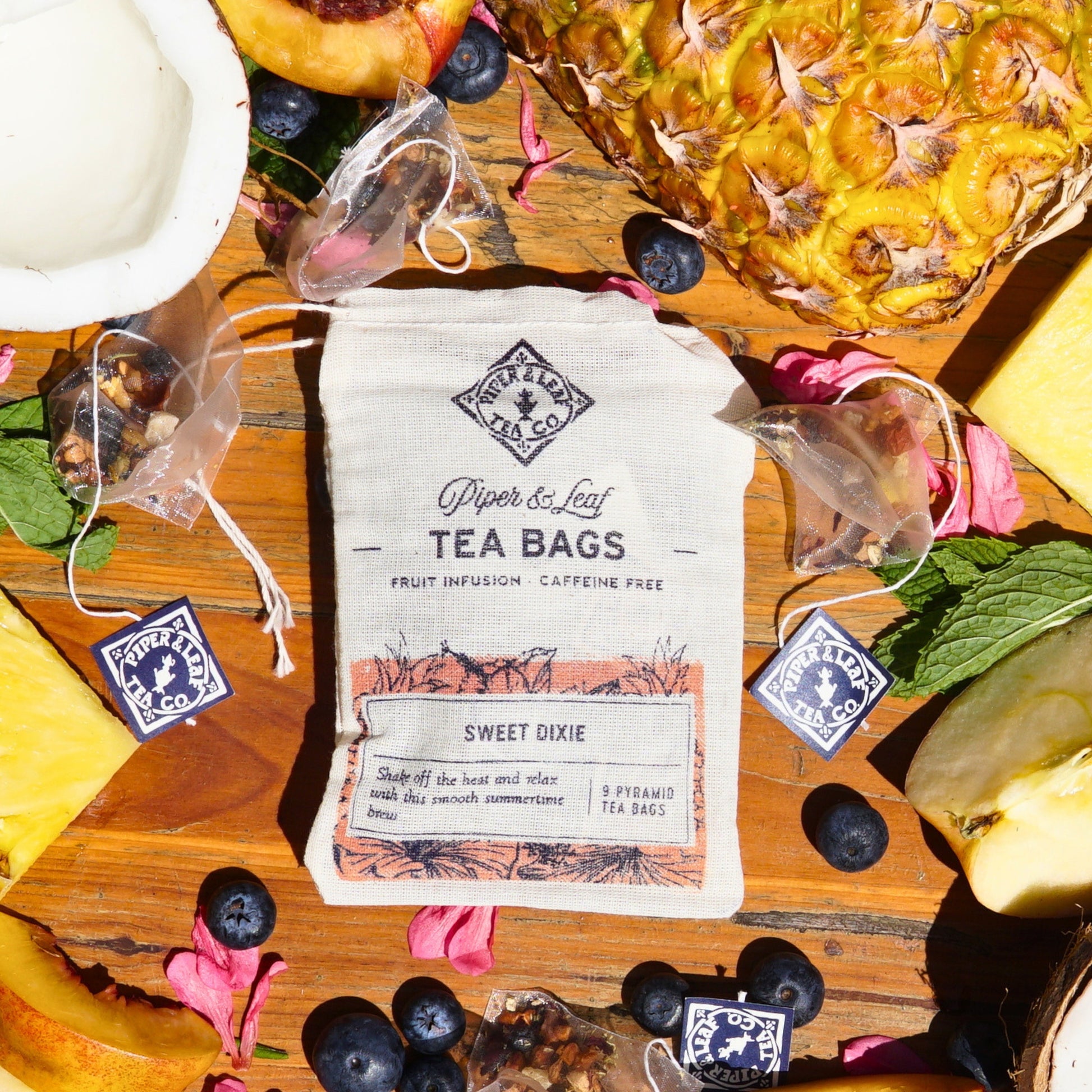 A package of Piper & Leaf Tea Co.'s "Sweet Dixie 9ct Tea Bags in Muslin" is surrounded by pineapple slices, blueberries, and flower petals on a wooden surface, creating a delightful summertime brew.