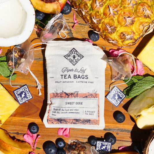 A package of Piper & Leaf Tea Co.'s "Sweet Dixie 9ct Tea Bags in Muslin" is surrounded by pineapple slices, blueberries, and flower petals on a wooden surface, creating a delightful summertime brew.
