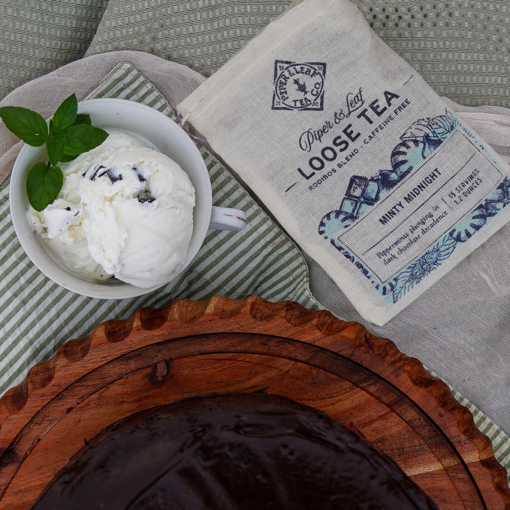 A cup of vanilla ice cream with mint leaves sits beside a Piper & Leaf Tea Co. Minty Midnight Muslin Bag of Loose Leaf Tea (15 Servings) and a rich chocolate cake on a wooden tray.