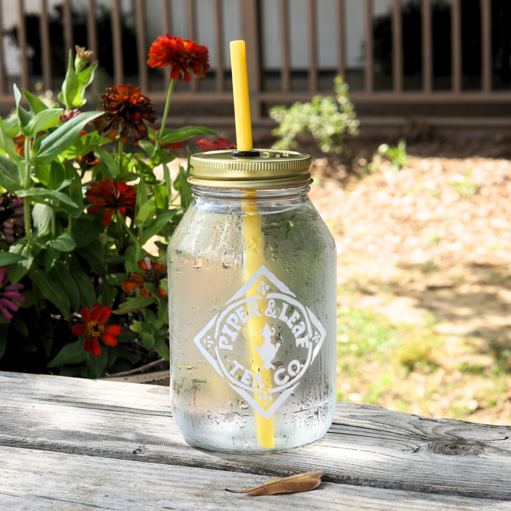 An UnStraw by Piper & Leaf Tea Co., consisting of a glass mason jar with a BPA-free yellow straw, is placed on a wooden table outdoors near flowers.