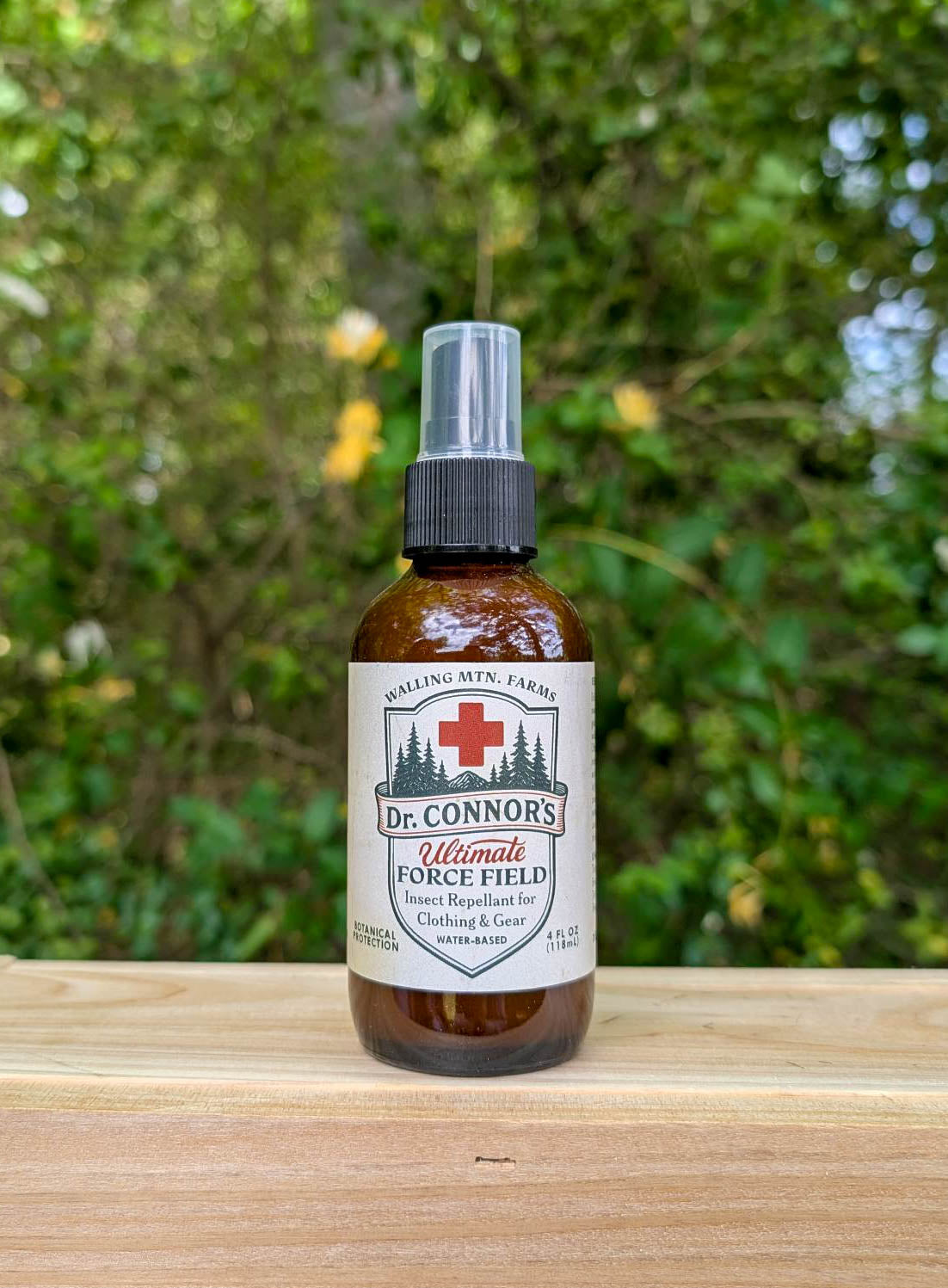 A brown bottle of Walling Mountain Farms' Dr. Connor's Ultimate Force Field Bug Spray for Clothing & Gear sits on a wooden surface with green foliage in the background.