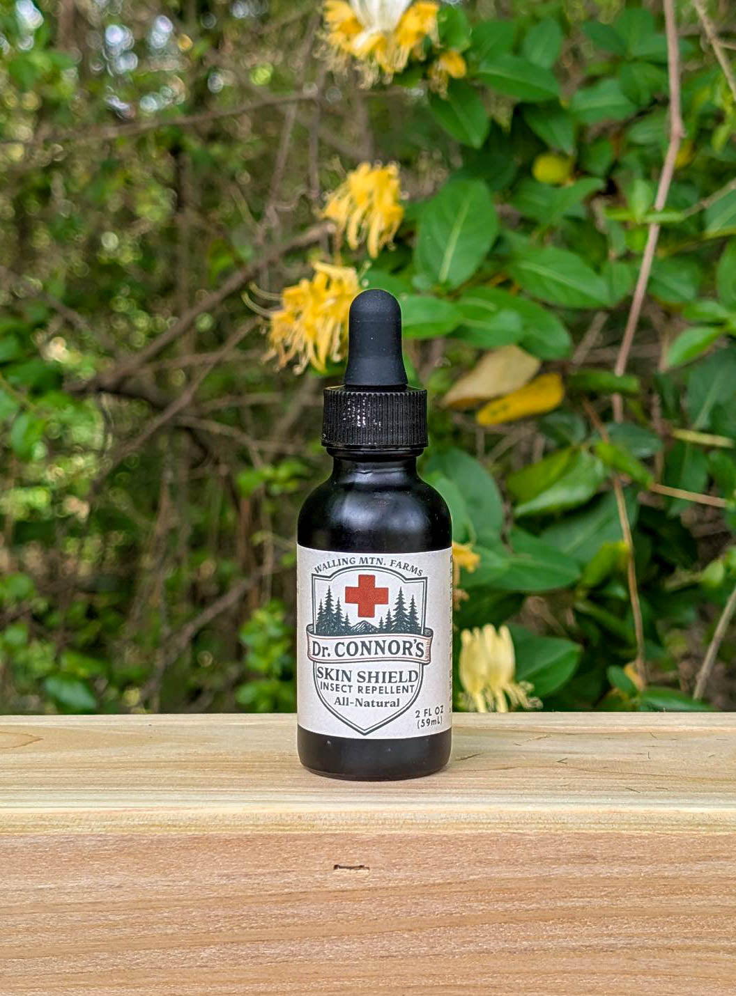 A small dropper bottle labeled "Dr. Connor's Skin Shield Insect Repellent - Rub-On Formula" from Walling Mountain Farms, a DEET-free, all-natural repellent, sits on a wooden surface with green foliage and yellow flowers in the background.