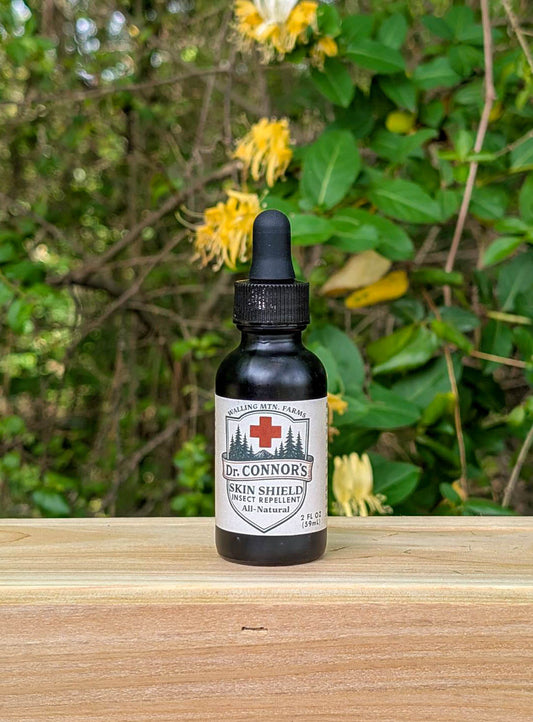 A small dropper bottle labeled "Dr. Connor's Skin Shield Insect Repellent - Rub-On Formula" from Walling Mountain Farms, a DEET-free, all-natural repellent, sits on a wooden surface with green foliage and yellow flowers in the background.
